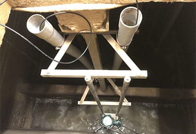 Two 3-inch wide white PVC pipes, each with a black cable, extend down near a pump in brown water ten or more feet below vantage point of picture