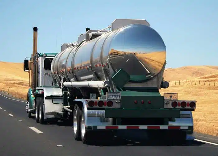 A metallic oil tank being pulled by a green semi-truck is driving through the desert, used to convey the idea of API 18.2, or the Custody Transfer of Crude Oil from Lease Tanks using Alternative Measurement Methods.